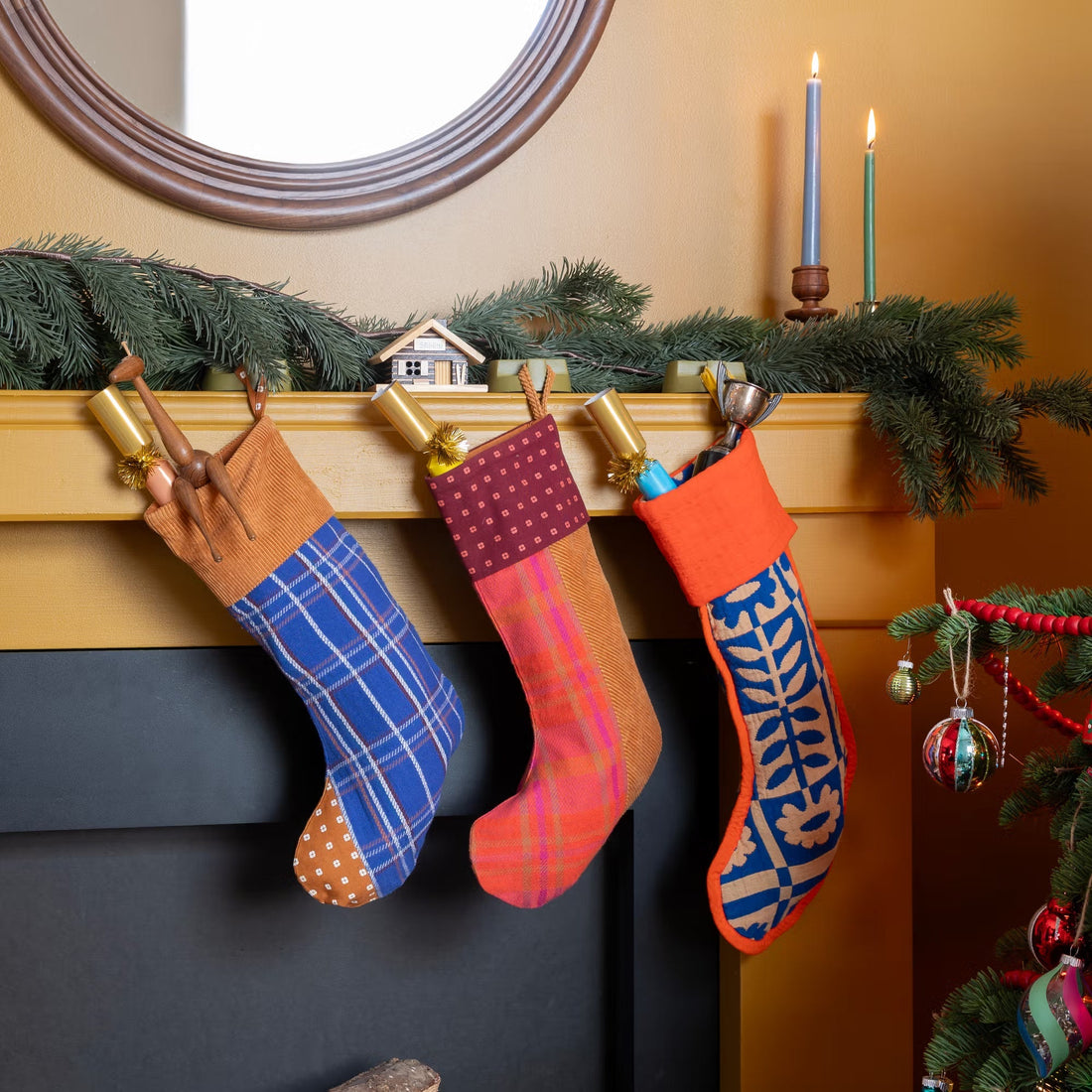 Holiday mantel with three colorful stockings, evergreen garland, candles, and Christmas tree ornaments