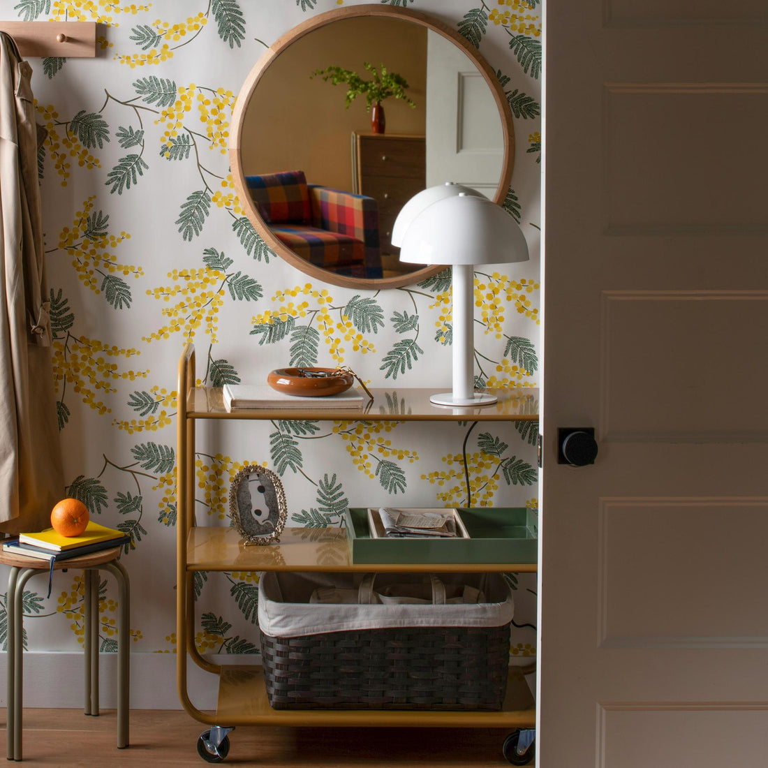 Round mirror hangs above a console in a floral-wallpapered entryway with a lamp and colorful chair reflected