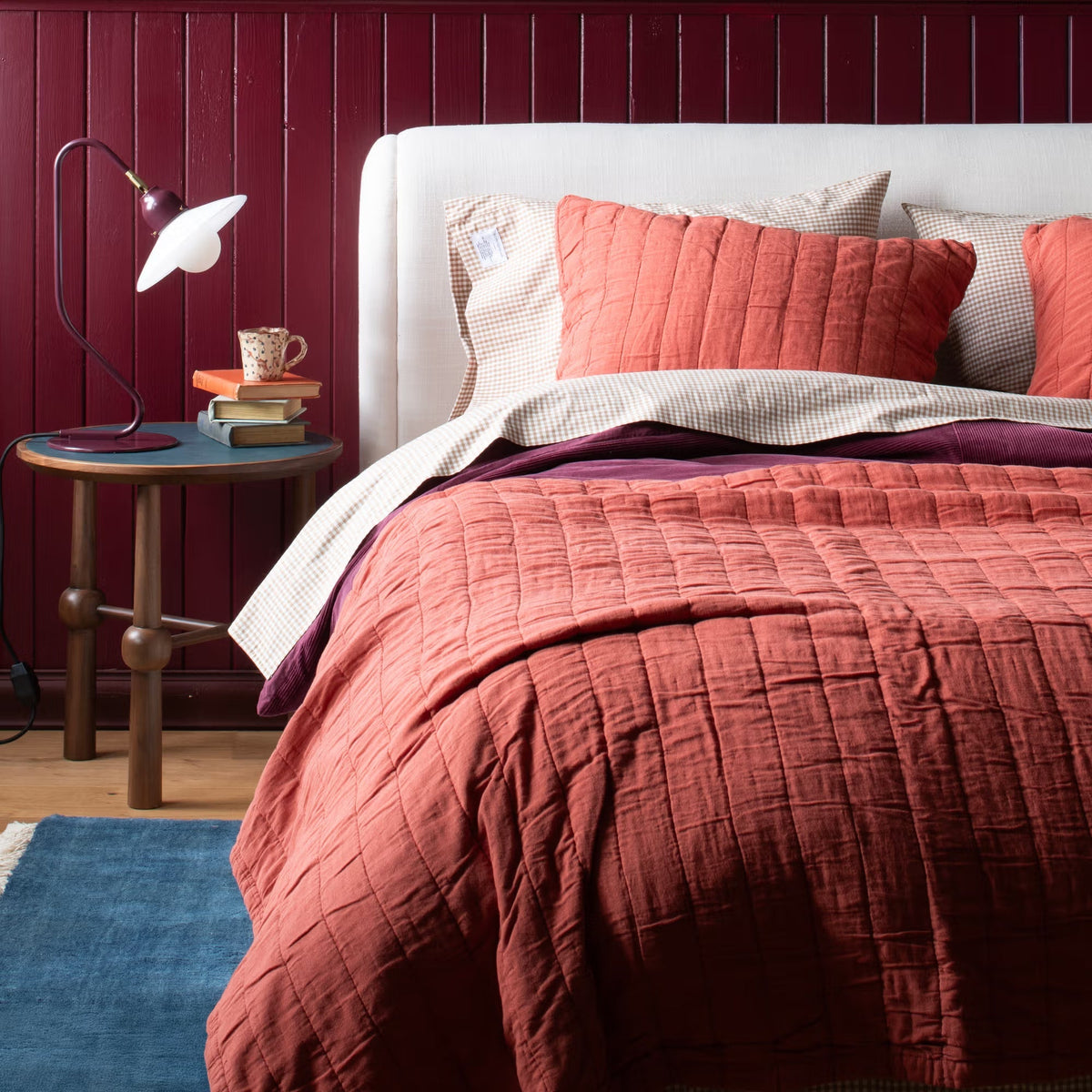 A bed layered with coral quilted bedding and gingham pillows beside a lamp and books