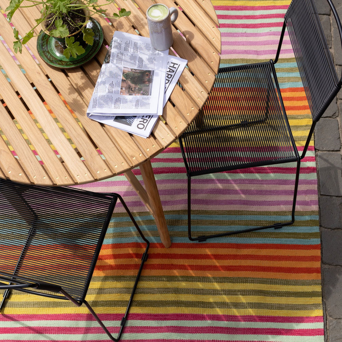 Outdoor table and chairs arranged on a colorful striped rug with a plant, coffee, and newspaper