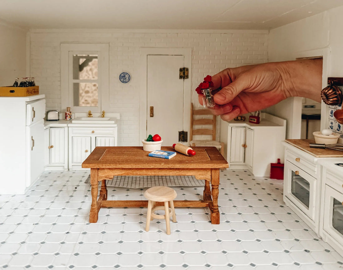 Hand holding a tiny vase of red flowers in a miniature dollhouse kitchen with white cabinets and a wooden table