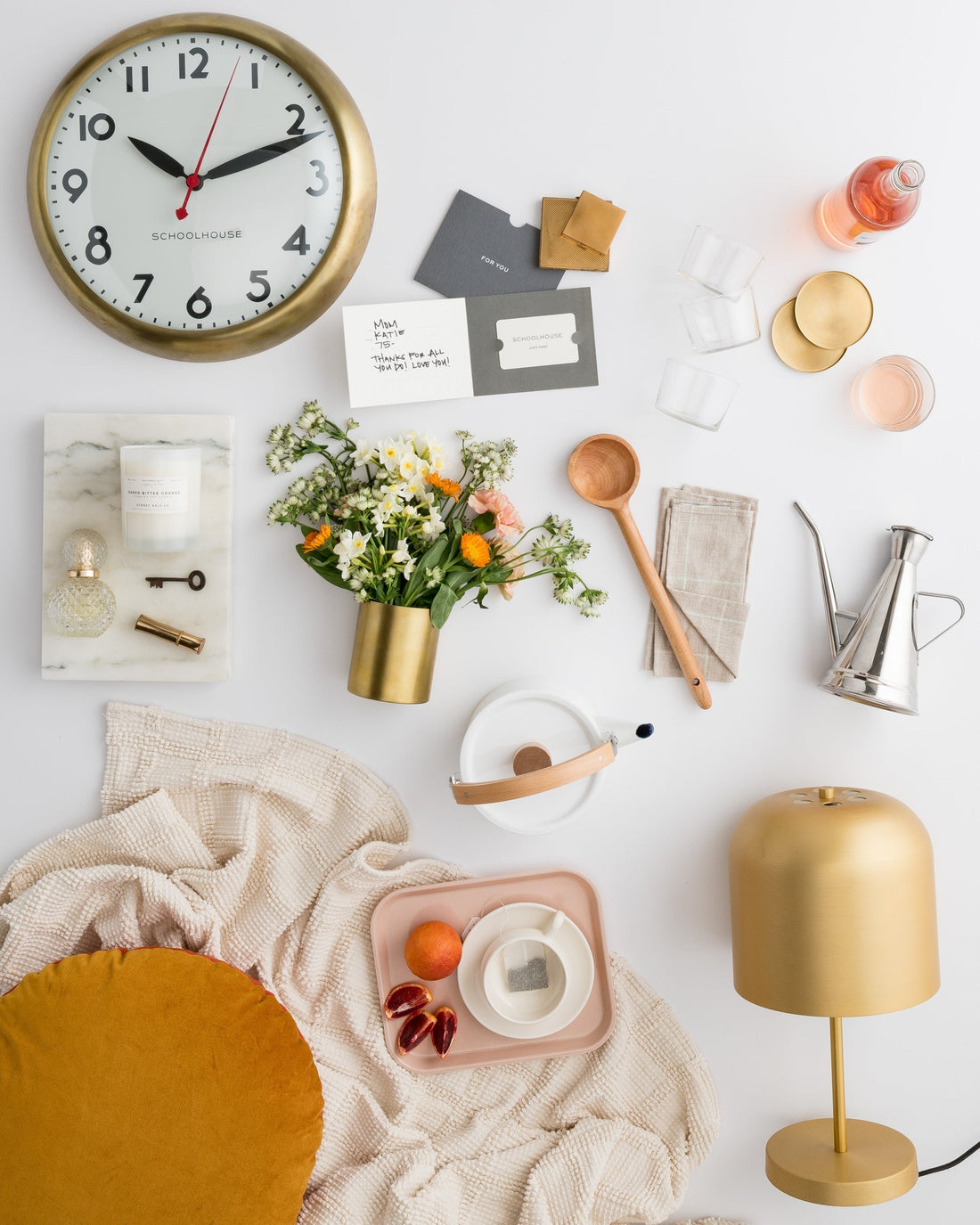 Flat lay of Mother’s Day gifts on white: Schoolhouse brass wall clock, bouquet in gold vase, wooden spoon