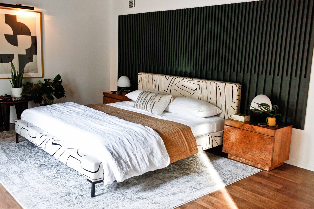 Sunlit master bedroom with patterned upholstered bed, white linens, brown throw, wood nightstands, black slatted accent wall and potted plants