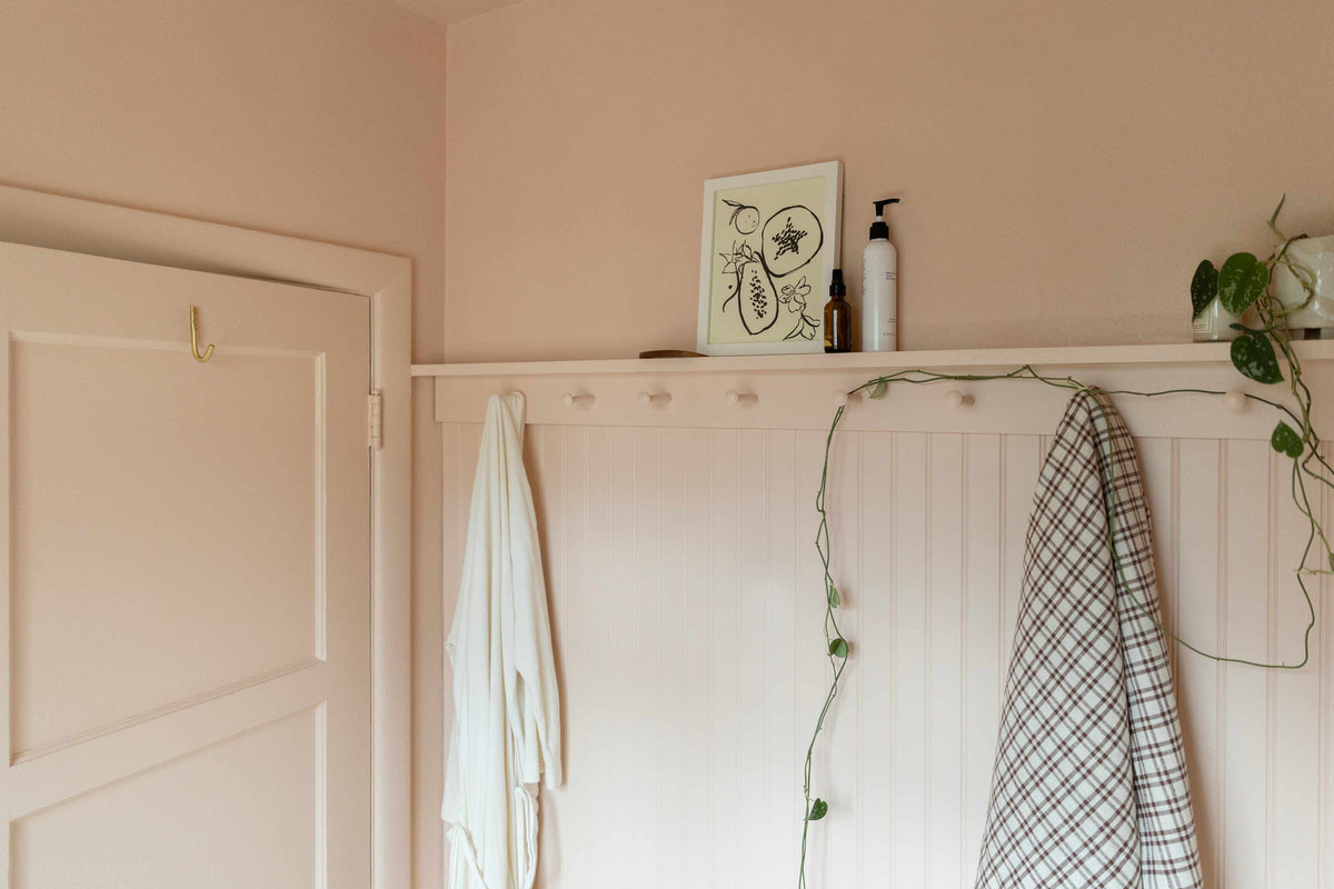 Pale pink beadboard wall in an entryway with a peg rail, a white robe and plaid towel hanging, framed artwork and a trailing pothos plant on a narrow shelf