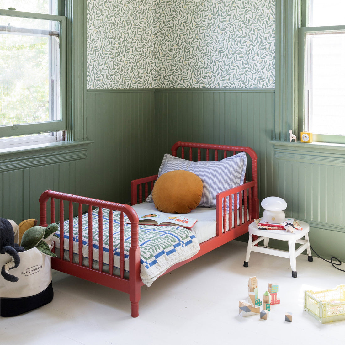 Red toddler bed with quilt and mustard pillow in green nursery with leafy wallpaper, white side table and wooden blocks