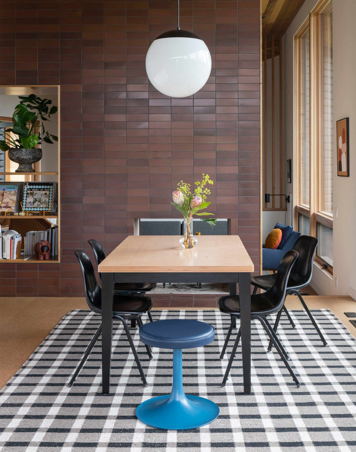 Dining table with vase of flowers, black chairs, plaid rug and round pendant light against a brown tiled accent wall in a sunlit dining area