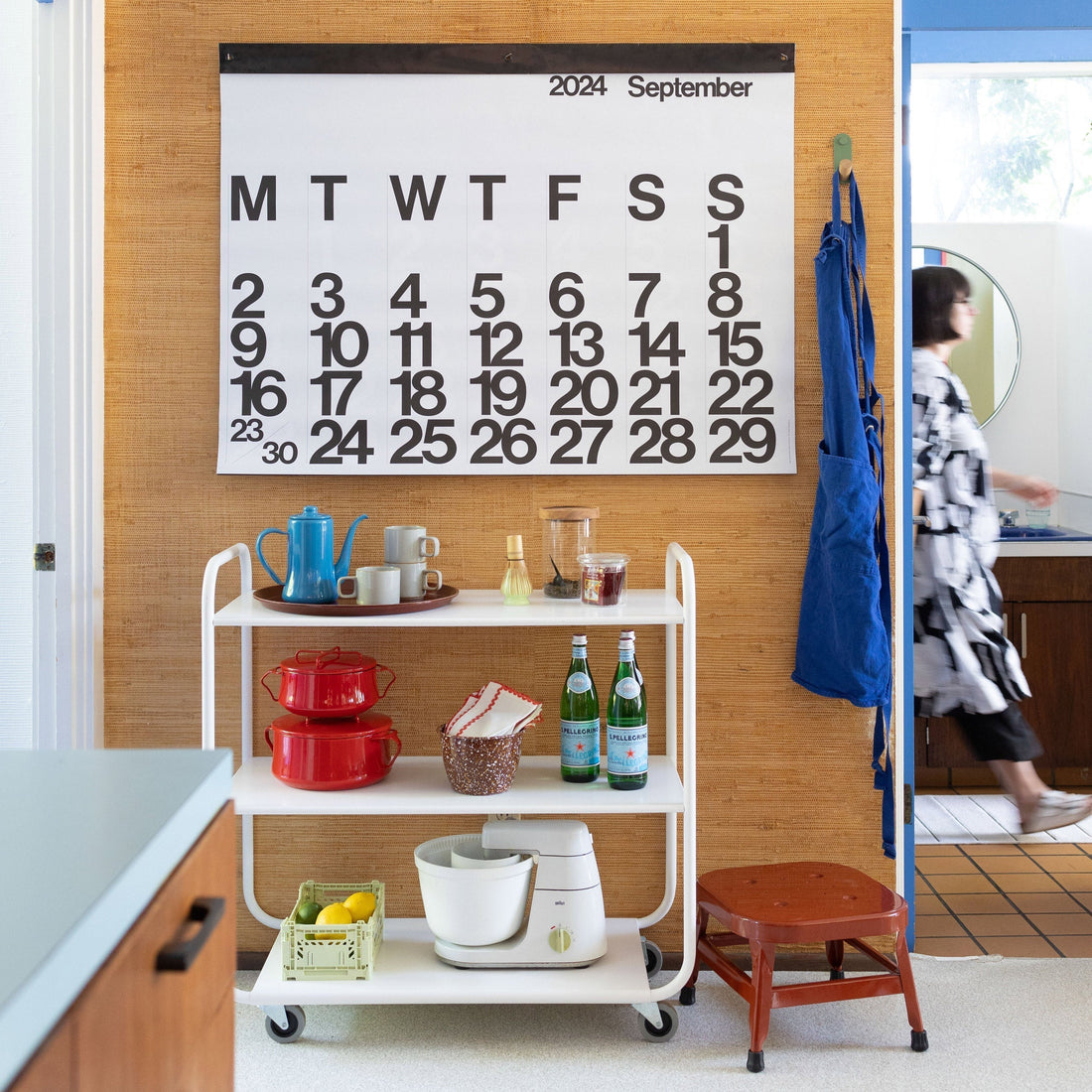 Cart with cookware, mugs and bottles under a calendar reading 2024 September; blue apron hanging and person walking by