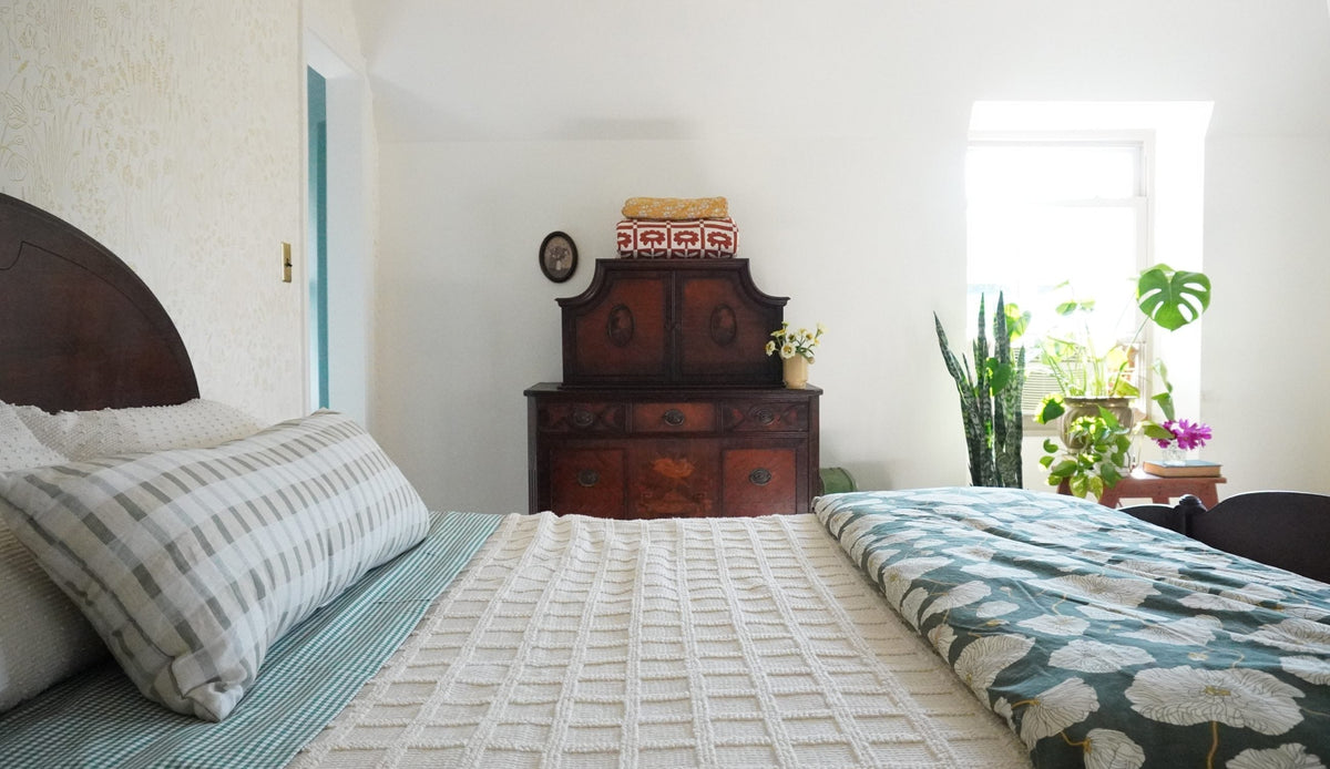 Bed and antique wooden dresser in a sunlit bedroom with potted plants by a window