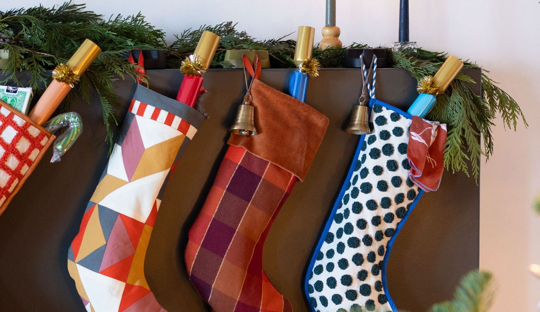 Colorful Christmas stockings hang from a dark mantel beneath evergreen garland and festive crackers