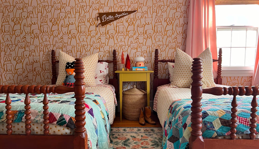 A bedroom with twin wooden beds, colorful quilts, dog-pattern wallpaper, pink curtains, and a yellow nightstand