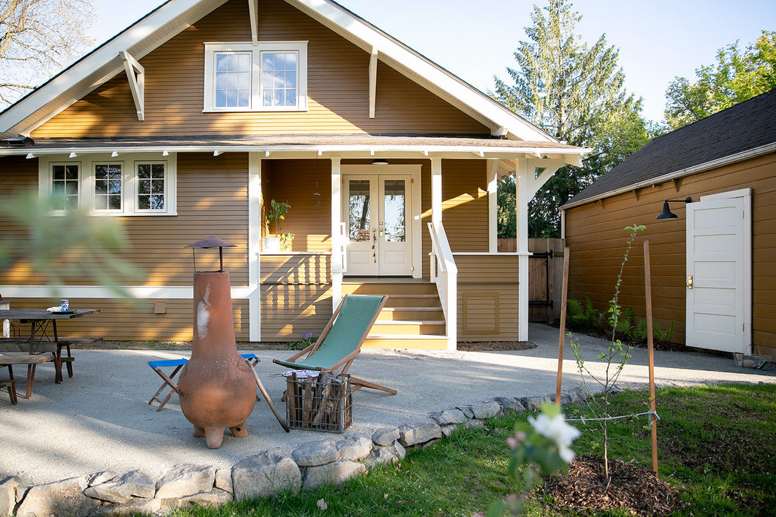 Sunlit front porch of a brown Craftsman house with white trim, patio chiminea, green canvas lounge chair, and detached shed