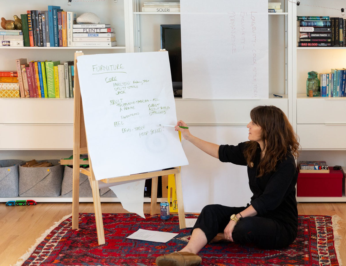 Woman seated on a rug pointing to an easel pad labeled Furniture in a bookshelf-lined office