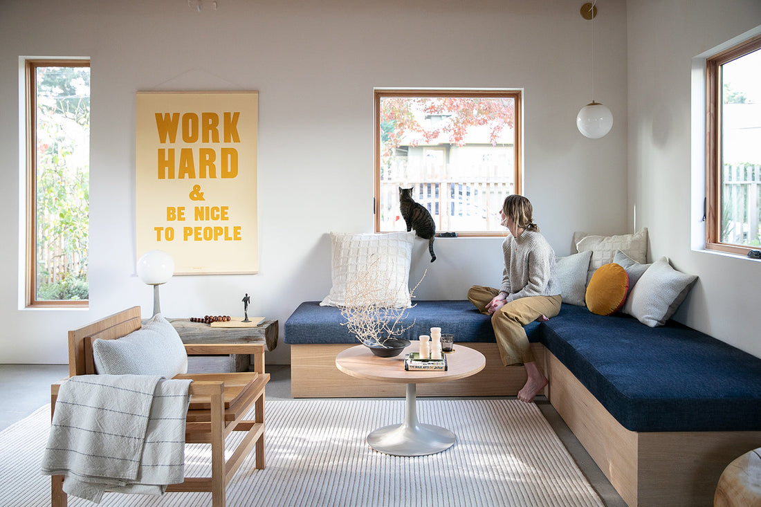 Woman on bench watching cat on windowsill in living room, yellow poster reads WORK HARD AND BE NICE TO PEOPLE