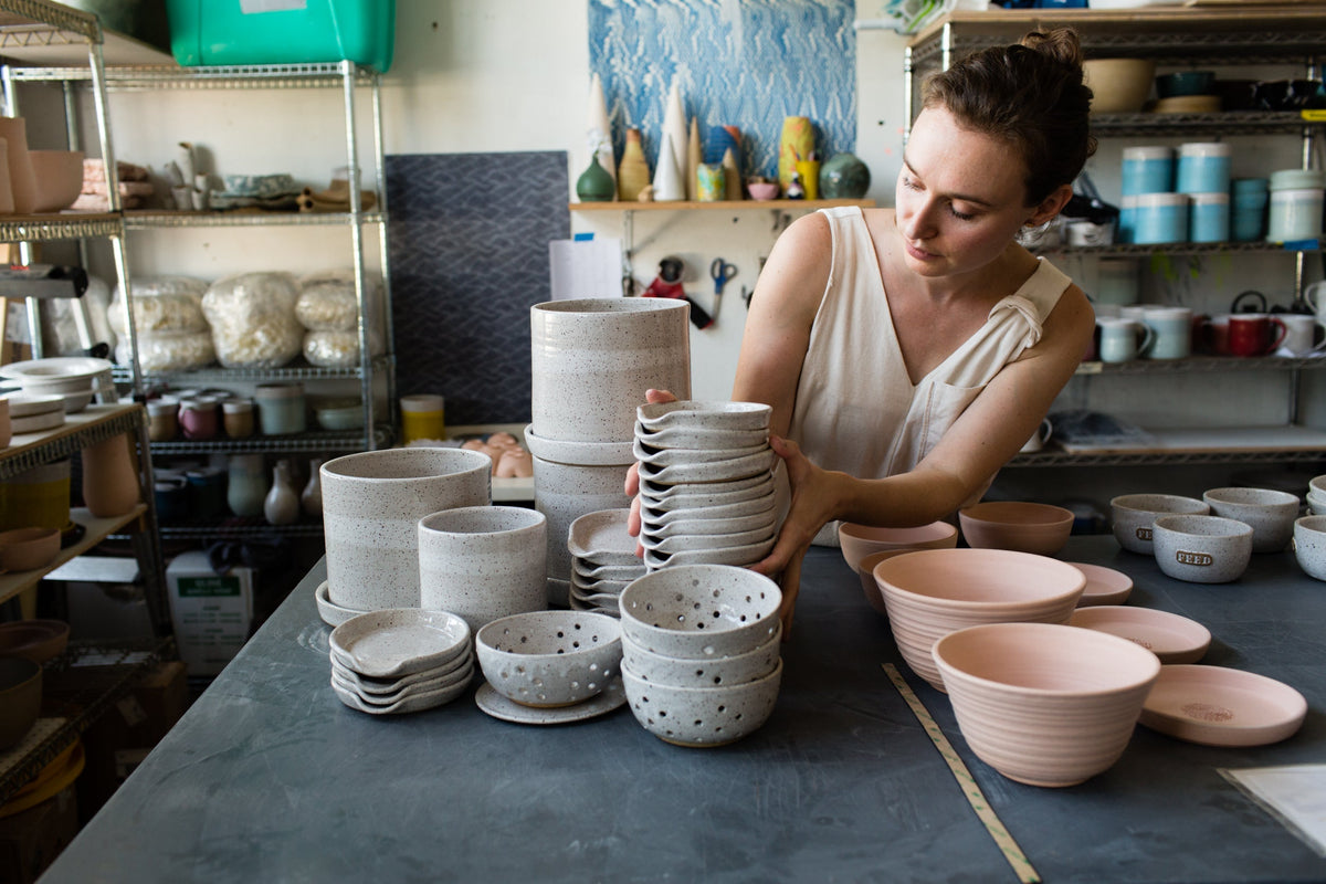 A potter arranging stacks of speckled stoneware bowls and pink ceramic bowls on a studio table with shelves of glazes behind her