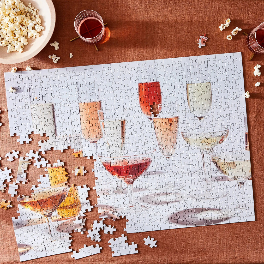 Jigsaw puzzle spread across a rust-colored table with cocktail glasses, popcorn, and loose pieces