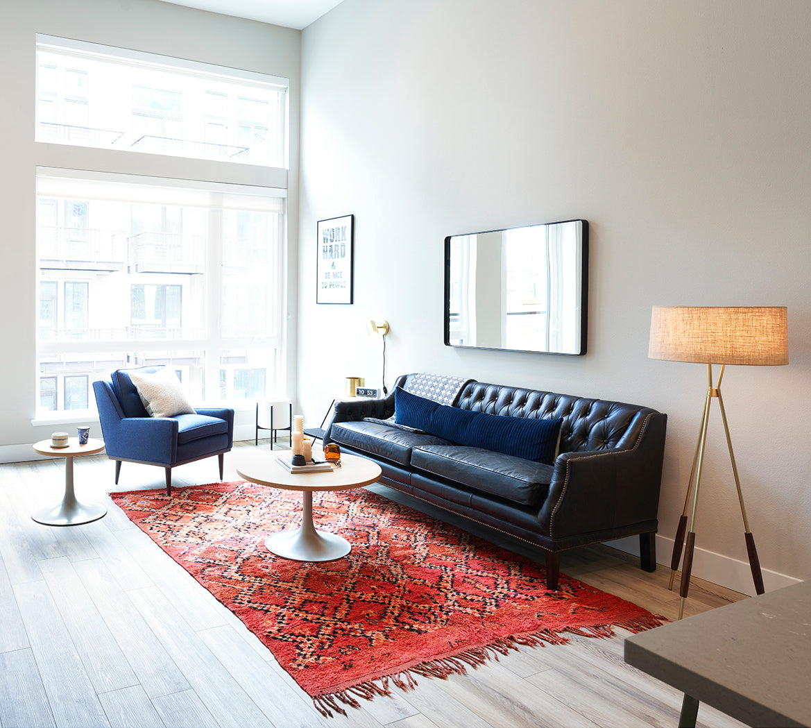 Sunlit living room with black tufted leather sofa, blue armchair, red patterned rug, round coffee tables, and tripod floor lamp