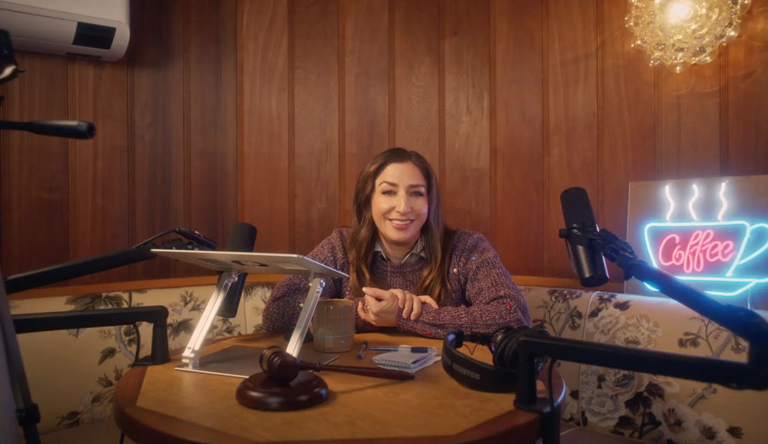 Chelsea Peretti sits at a table with microphones in a wood-paneled podcast studio with a neon Coffee sign