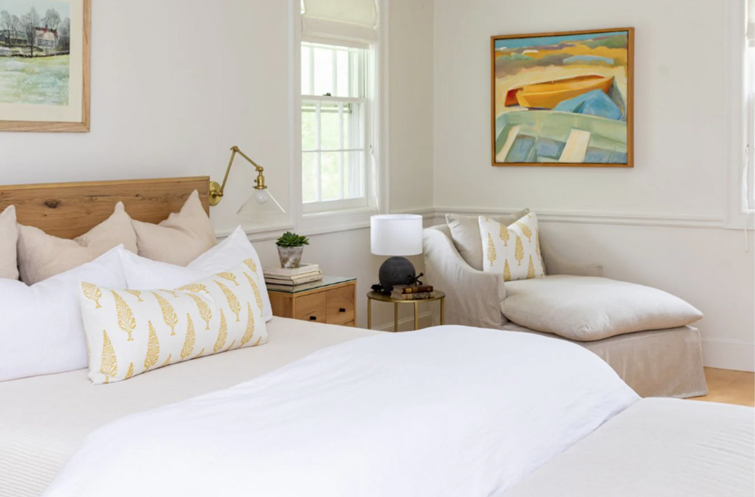 Bright bedroom with a white bed, gold botanical pillows, armchair, artwork, and natural light