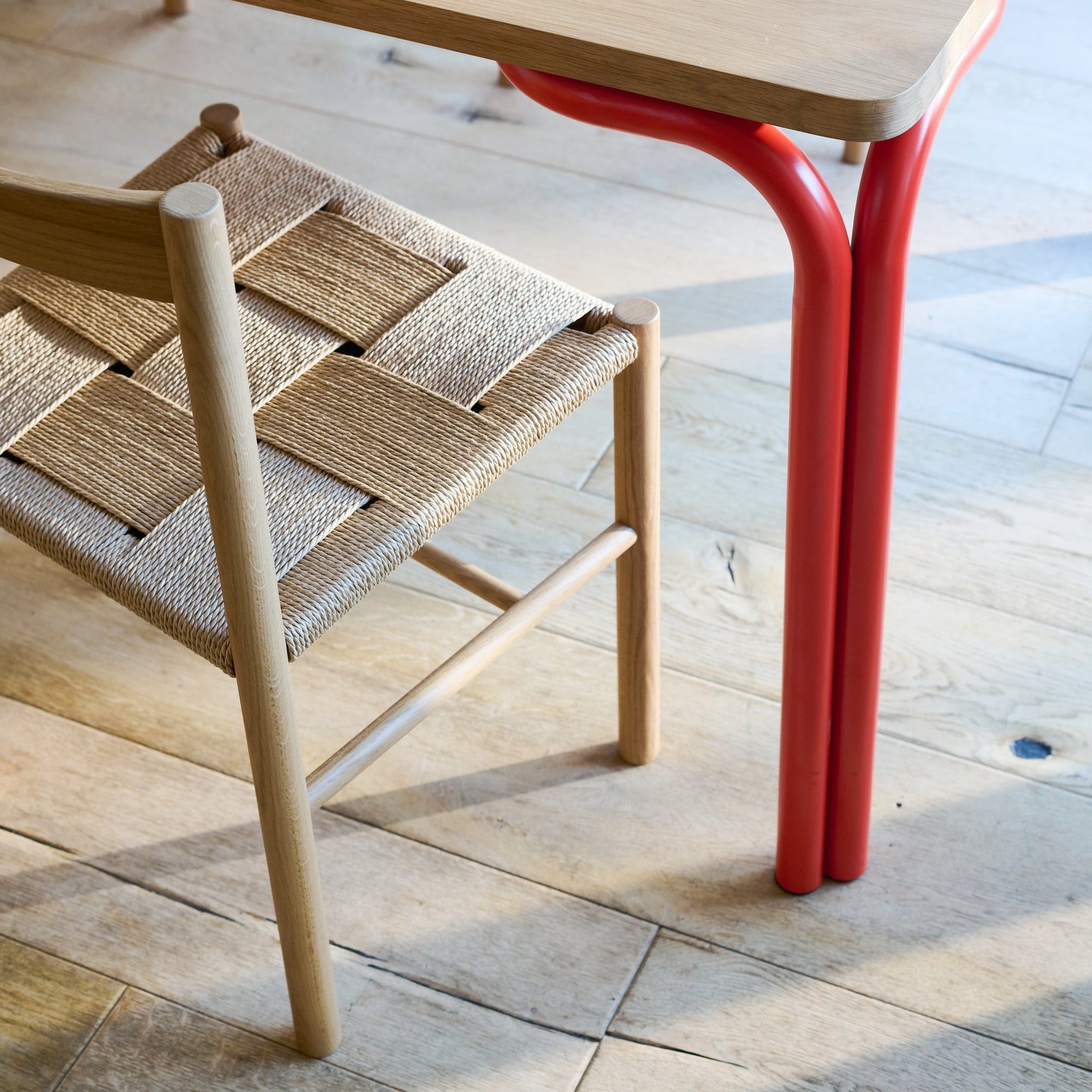 Close-up of Forma Dining Table corner with oak rounded top and red tubular legs next to woven oak chair