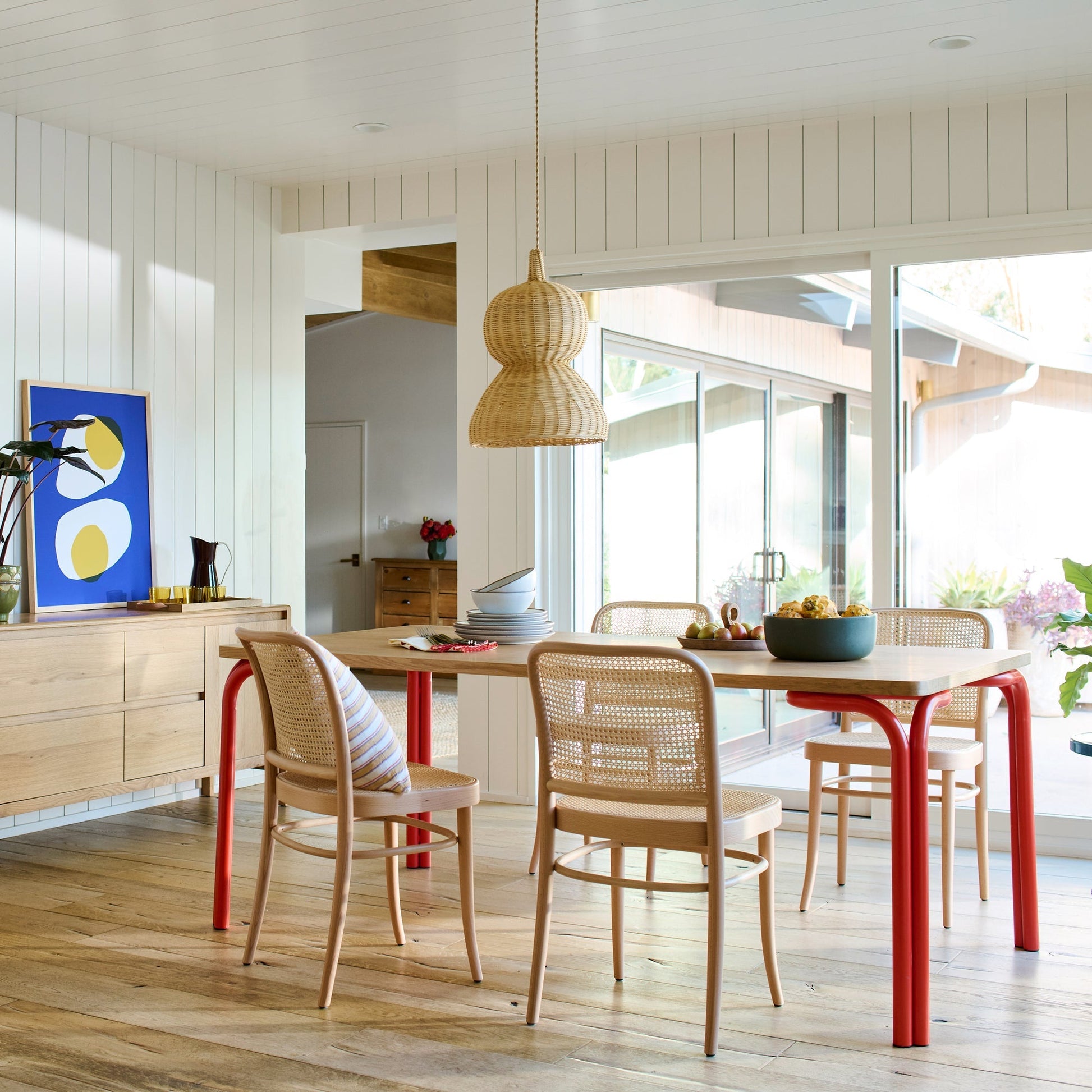 Three-quarter view of Forma dining table showing light wood top and bold red curved legs with cane-back chairs and a rattan pendant in a sunlit dining room