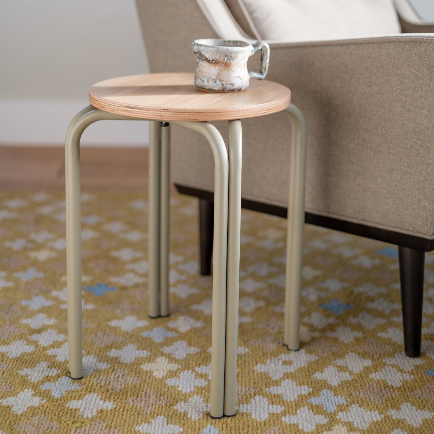 Close-up of Forma Stool showcasing white oak round top with exposed plywood edge and powder-coated steel legs beside an upholstered chair