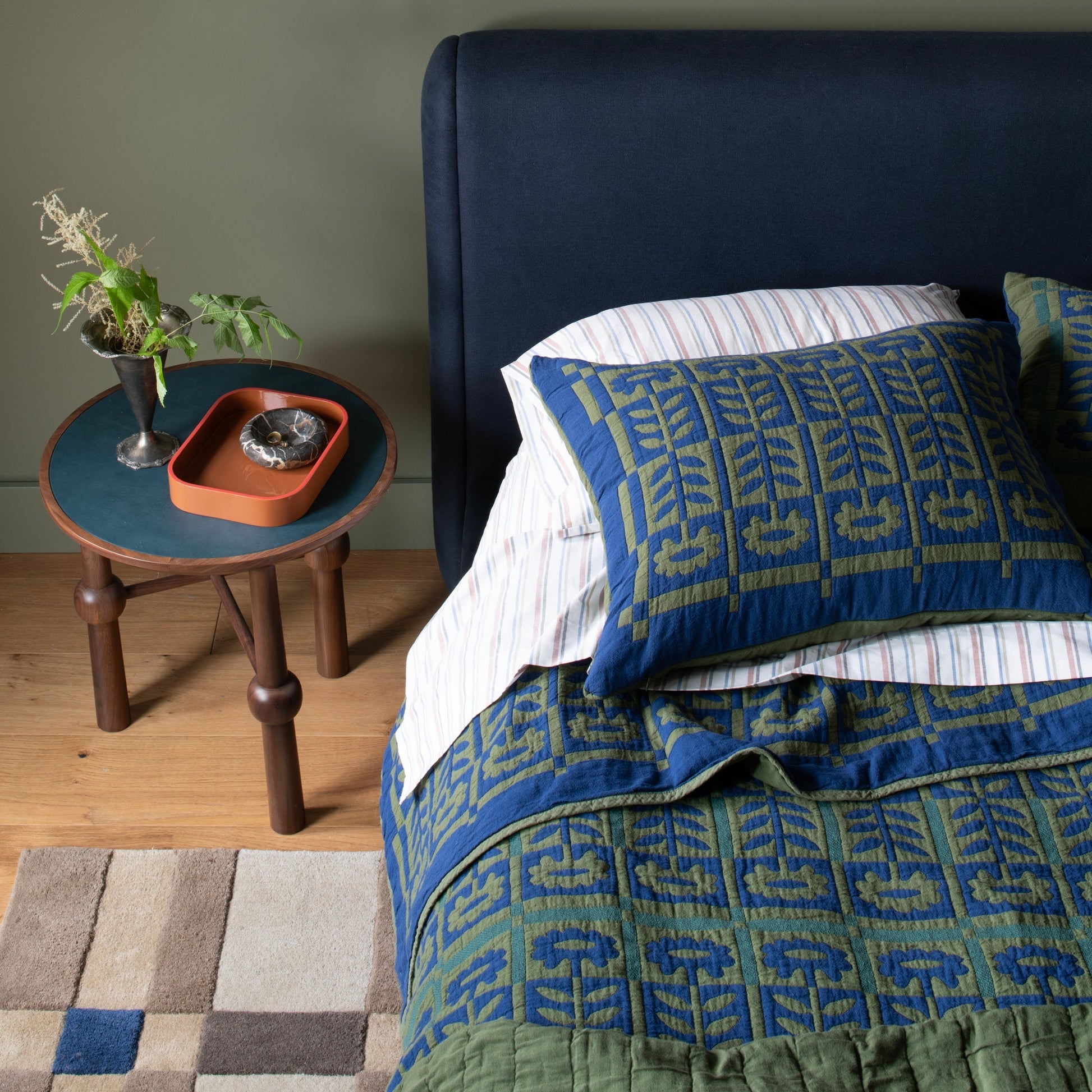 Ann Occasional Table beside a bed, styled with a metal vase of greenery, an orange tray, and a navy upholstered headboard