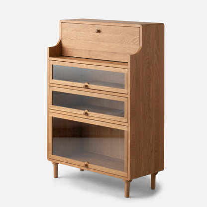 Three-quarter view of Peggy Cabinet showcasing a light wood finish, upper drawer, and three glass-front compartments with tapered legs