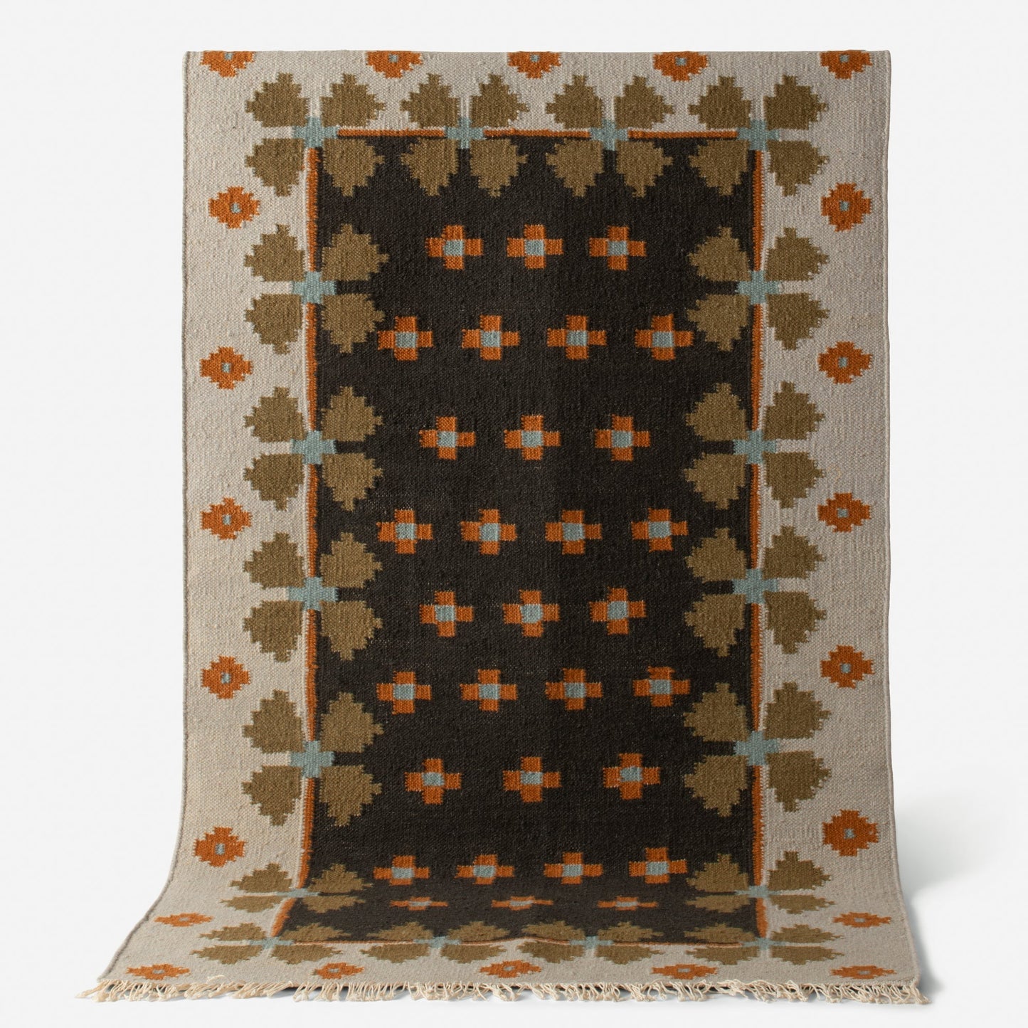 Angled view of Flora Wool Flatweave Rug showcasing geometric Scandinavian folk-floral pattern in black, tan, orange, and blue with fringed edge on white background