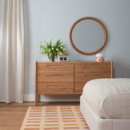 Terracotta Network Tufted Wool Rug lies beside a bed in a bedroom with a wood dresser, round mirror, and flowers