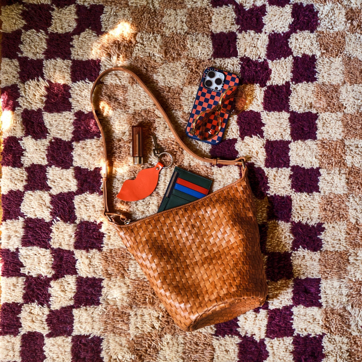 Top-down view of Schoolhouse x Clare V. Felix Shag Wool Rug in raisin and butterscotch checker pattern showcasing high-pile wool texture with a woven leather purse and phone