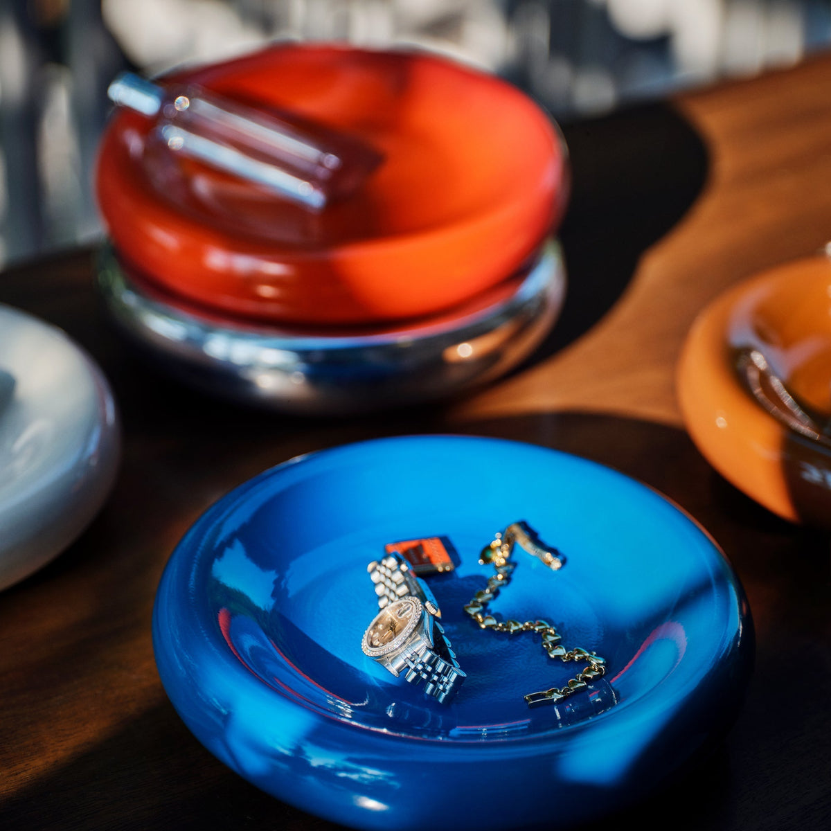 Cobalt-gloss Yves Cast Metal Catch-All holds a watch and necklace on a table with colorful catch-all dishes behind it