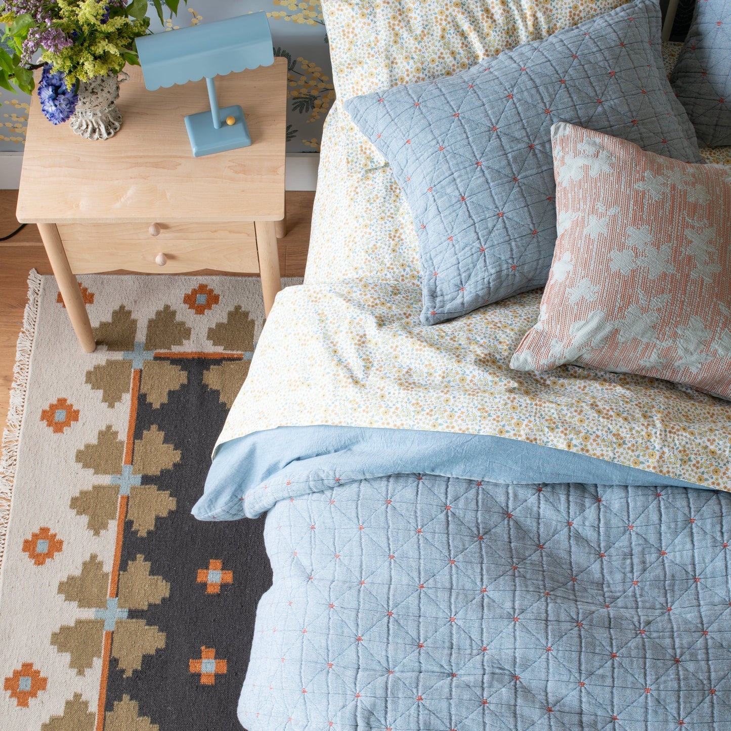 Flora Wool Flatweave Rug peeking from under a bed with a blue quilt, floral sheets, and a light wood nightstand with a lamp