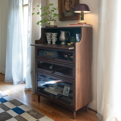 Sunlit corner with Campus Wool Rug on a hardwood floor beside a wooden side cabinet, potted plant, and table lamp