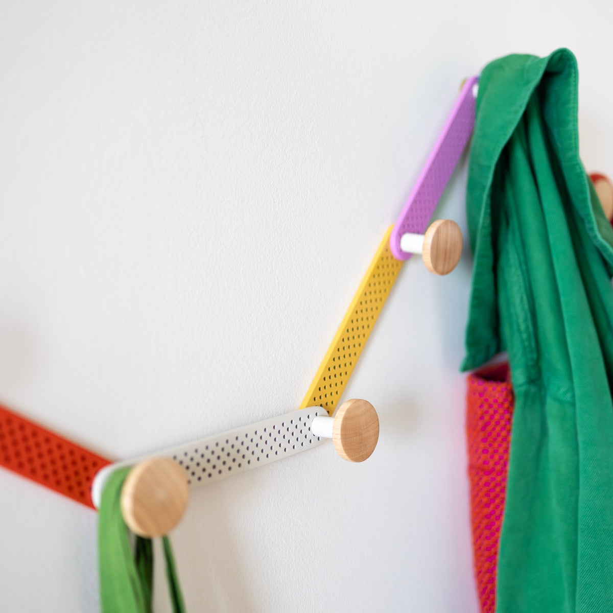 Close-up of Tula Hook showcasing perforated multicolor metal and natural wood finials with coats on a white wall