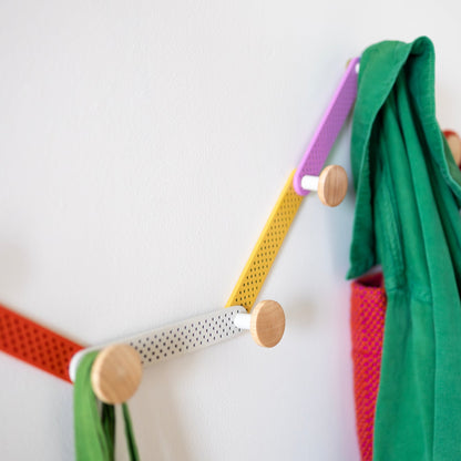 Close-up of Tula Hook showcasing perforated multicolor metal and natural wood finials with coats on a white wall