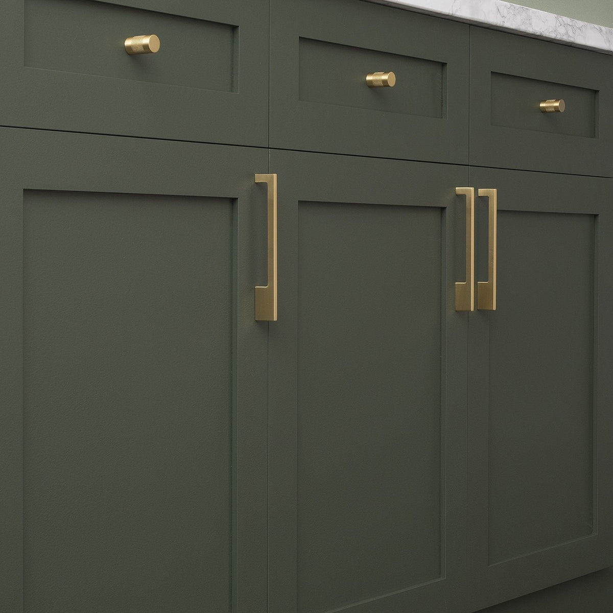 Front view of Alberta Pull showcasing its thin asymmetrical brass design installed on dark green cabinetry