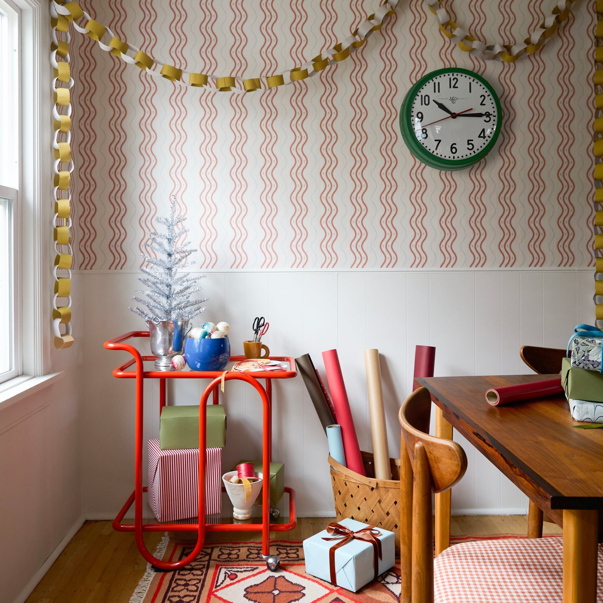 Dining nook styled with Linnea wool flatweave rug, red serving cart, wrapped gifts, patterned wallpaper, and green wall clock