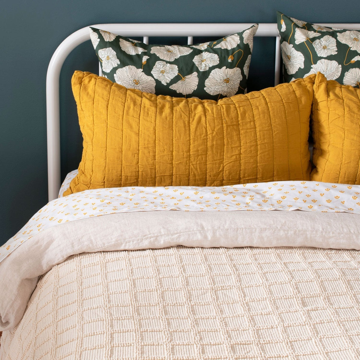 Yellow channeled cotton sham on a bed with floral pillows and white bedding against a dark blue wall