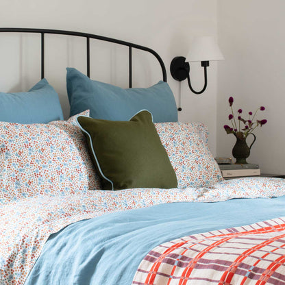 Angled view of Wallowa Floral Percale Sheet Set on a made bed showing small blue and rust floral print, blue pillowcases, and olive green accent pillow