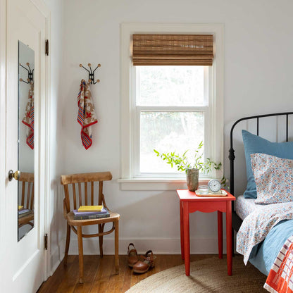 Bed dressed in Wallowa Floral percale sheets beside a window with blue pillow, red bedside table, plant and wooden chair