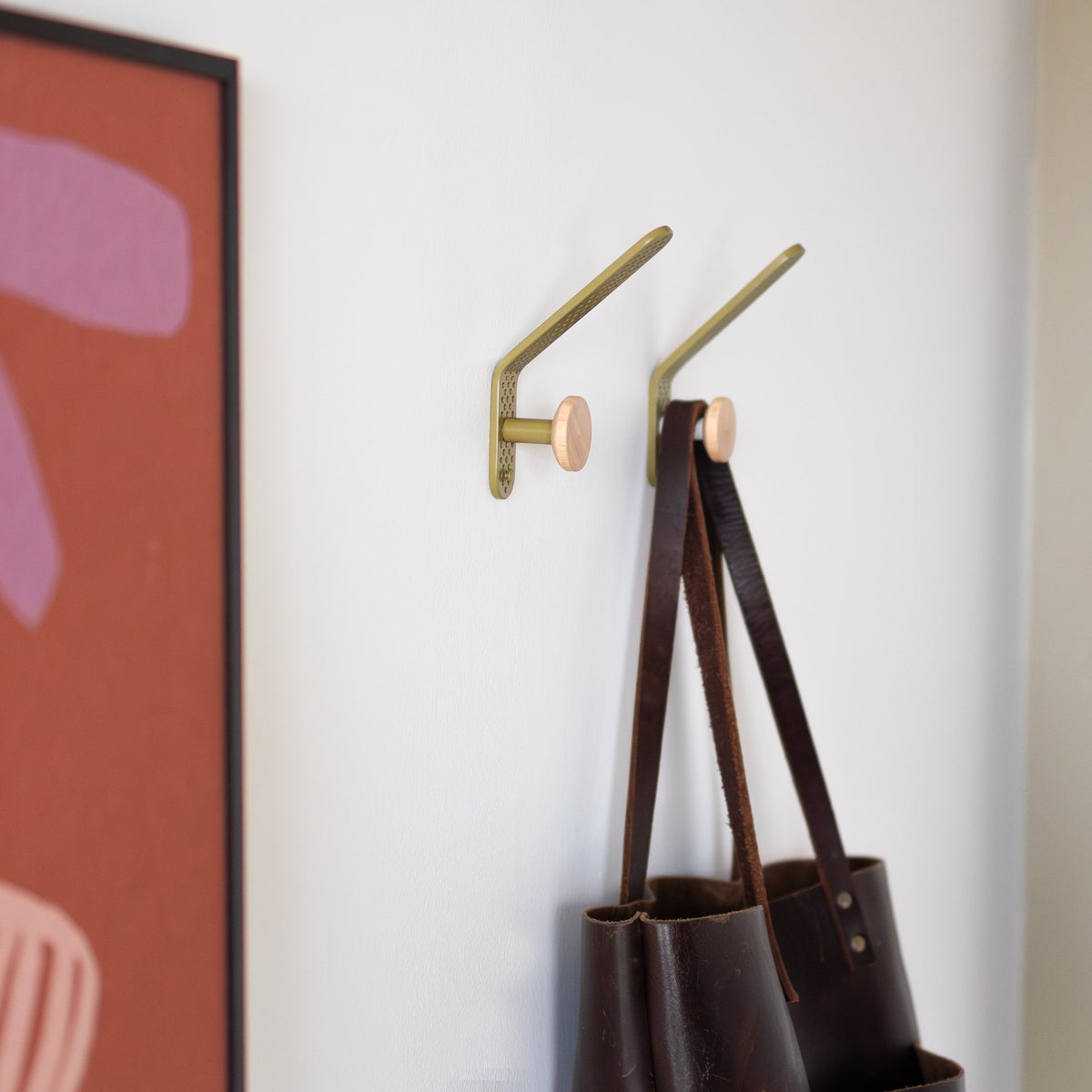 Two olive satin Tula hooks hold bags on a white wall beside colorful artwork