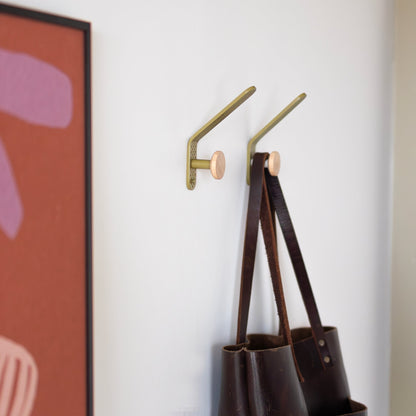 Two olive satin Tula hooks hold bags on a white wall beside colorful artwork