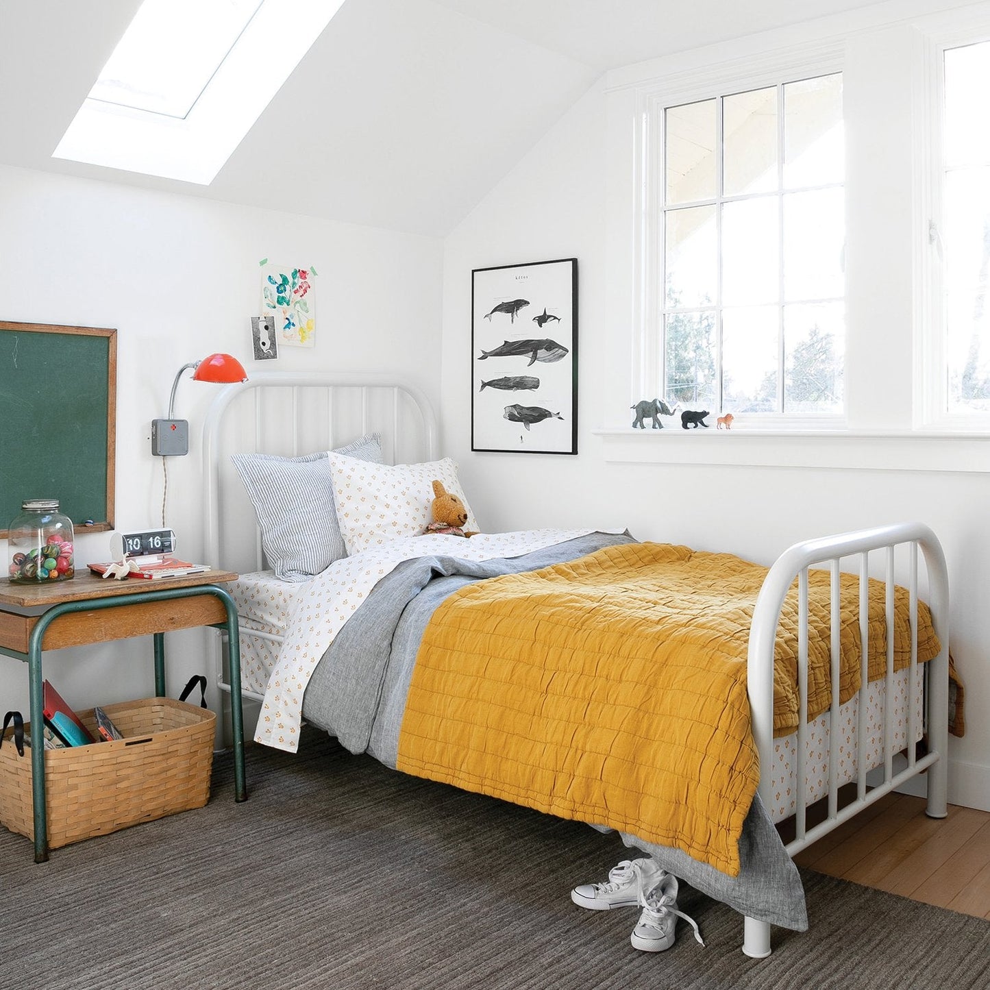 Single white metal bed styled with Diamond Ticking Quilted Pillow Sham, gray sheets and mustard quilt in bright bedroom