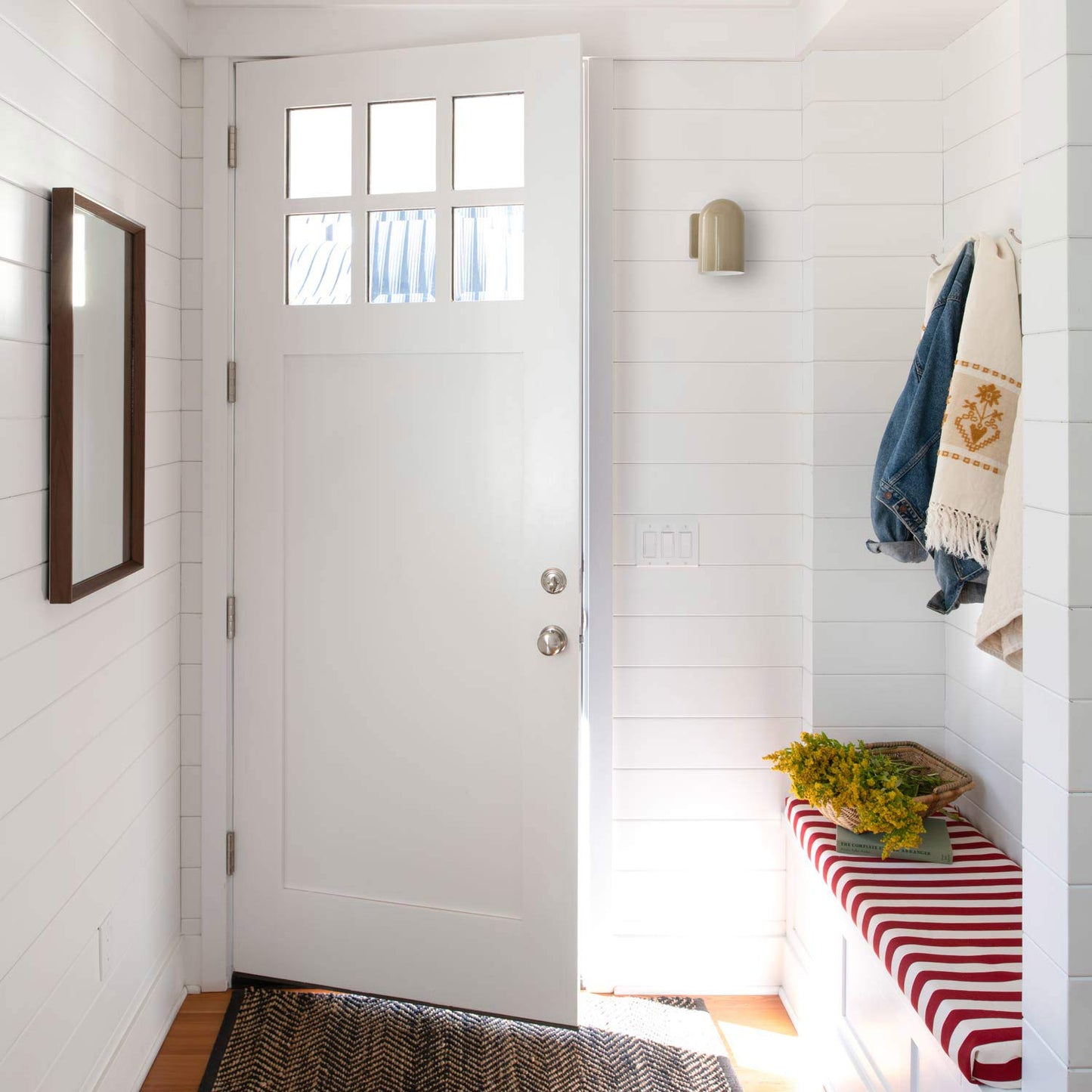 Entryway with open white door and Allegheny lichen gloss wall sconce mounted on white shiplap above a red-and-white striped bench with a denim jacket and basket of yellow flowers