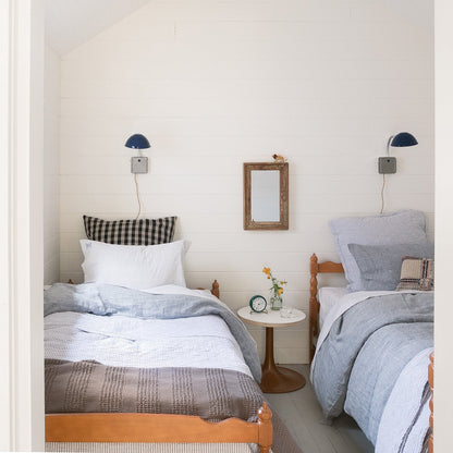 Two twin beds with gray diamond-ticking quilted pillow shams and blue-gray linen bedding in a bright white shiplap bedroom