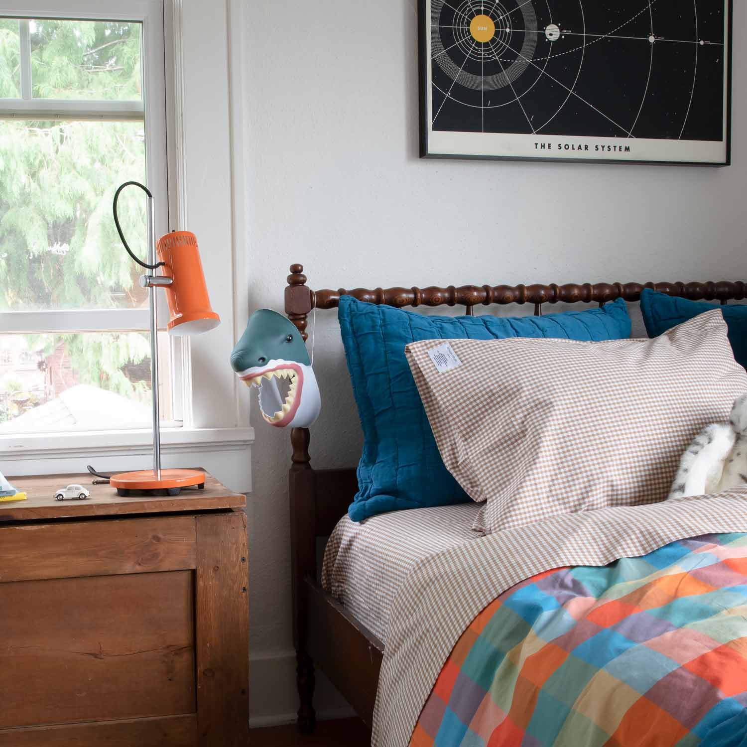 Gingham pillowcases on bed with teal pillows, orange bedside lamp, and framed print reading THE SOLAR SYSTEM