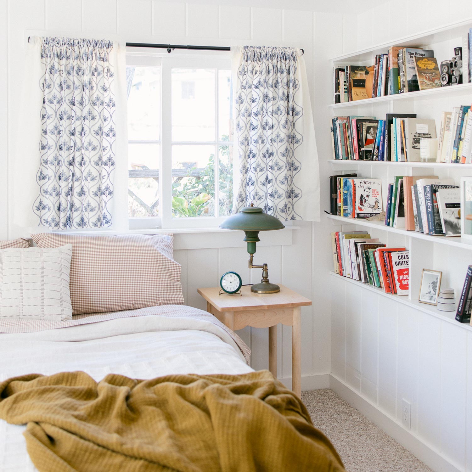Bed with russet gingham pillowcases beside a wooden nightstand and bookshelf in a bright cozy bedroom