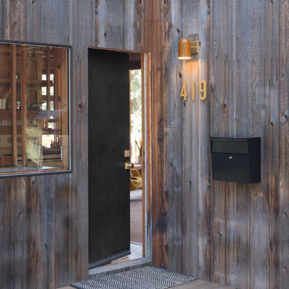 Mounted view of Allegheny 4.5-inch indoor outdoor sconce in brass, casting light on weathered wood by open black door