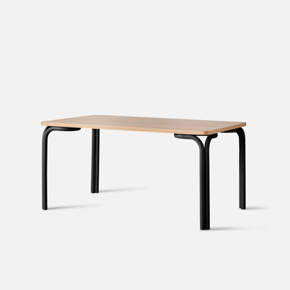 Three-quarter view of Forma Dining Table showing butterscotch satin wood top with rounded edges and curved matte-black tubular legs on a white studio background
